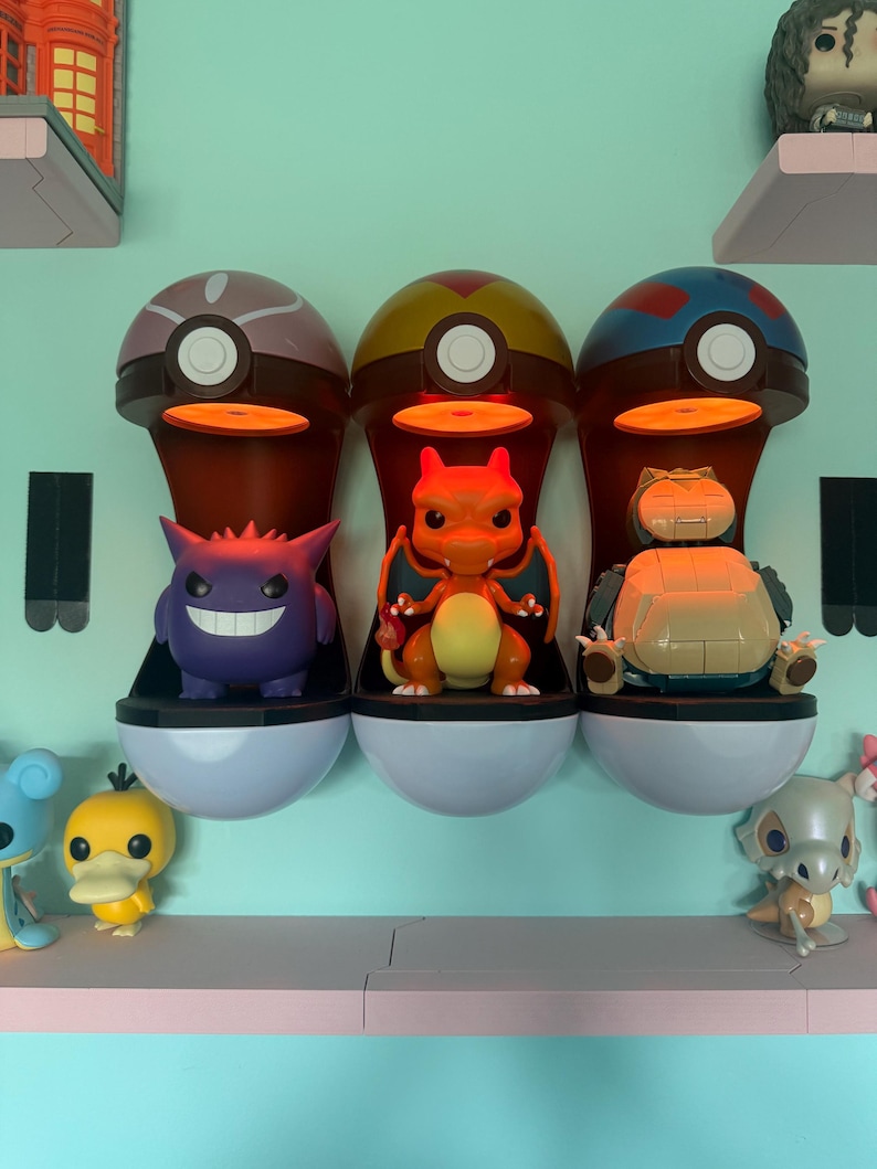 Op de afbeelding: Drie Pok&eacute;mon-thema displayplanken, elk met een ander kleurenschema, bevatten karakterbeeldjes. De planken zijn gevormd als Pok&eacute; Balls, met een licht dat van bovenaf schijnt. De beeldjes zijn onder andere Gengar, Charizard en Snorlax.