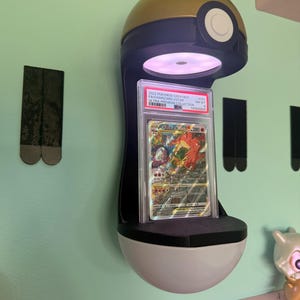 May include: A wall-mounted display stand shaped like a gold, blue, and white Pok&eacute; Ball featuring an integrated light that illuminates a graded Charizard VSTAR trading card encased in a clear plastic PSA slab with a white label at the top.