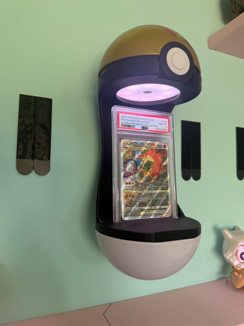 May include: A wall-mounted display stand shaped like a gold, blue, and white Pok&eacute; Ball featuring an integrated light that illuminates a graded Charizard VSTAR trading card encased in a clear plastic PSA slab with a white label at the top.