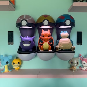 May include: Three wall-mounted, illuminated Pokeball display cases, each housing a vinyl figure. The left case holds a purple Gengar, the center case contains an orange Charizard, and the right case features a blue and cream-colored Snorlax figure.