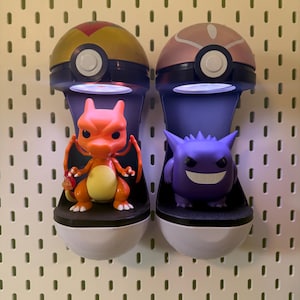 Op de afbeelding: Twee aan de muur gemonteerde display cases, elk in de vorm van een Pok&eacute; Ball, houden een Charizard en een Gengar figuur vast. De Charizard is oranje en geel, terwijl de Gengar paars is. De cases hebben een wit en zwart basis.