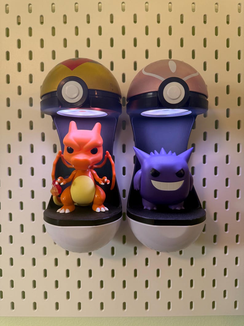 Op de afbeelding: Twee aan de muur gemonteerde display cases, elk in de vorm van een Pok&eacute; Ball, houden een Charizard en een Gengar figuur vast. De Charizard is oranje en geel, terwijl de Gengar paars is. De cases hebben een wit en zwart basis.