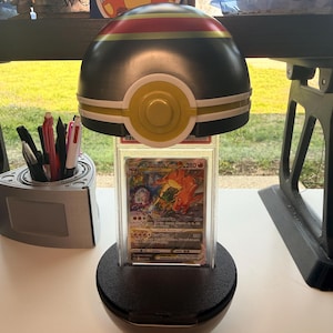 Puede incluir: Un expositor negro y dorado con forma de Poké Ball que sostiene una carta coleccionable de Charizard calificada. El expositor tiene una base circular y una parte superior en forma de cúpula con rayas rojas y verdes. La tarjeta está dentro de un soporte de plástico transparente.