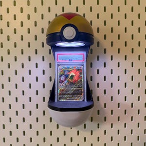 May include: A wall-mounted display light shaped like a gold, blue, and white Pok&eacute; Ball. The device features an integrated LED light that illuminates a vertically oriented, PSA-graded Charizard VSTAR trading card encased in a clear plastic slab.