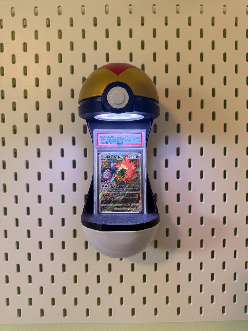 May include: A wall-mounted display light shaped like a gold, blue, and white Pok&eacute; Ball. The device features an integrated LED light that illuminates a vertically oriented, PSA-graded Charizard VSTAR trading card encased in a clear plastic slab.