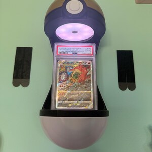 May include: A Pokémon card in a protective case, displayed in a Poké Ball-shaped holder. The card features a Charizard VSTAR illustration. The holder is black, white, and gold, with a built-in light. The card is graded and labeled with details.