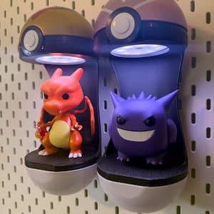 Op de afbeelding: Twee verlichte display stands in de vorm van Pok&eacute; Balls, elk met een Pok&eacute;mon figuur. De ene stand toont een Charizard, de andere een Gengar. De stands zijn wit, zwart en grijs, gemonteerd op een wit pegboard.