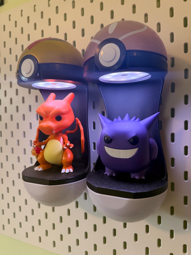 Op de afbeelding: Twee verlichte display stands in de vorm van Pok&eacute; Balls, elk met een Pok&eacute;mon figuur. De ene stand toont een Charizard, de andere een Gengar. De stands zijn wit, zwart en grijs, gemonteerd op een wit pegboard.