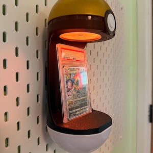 May include: A wall-mounted display stand shaped like a gold and white Pok&eacute; Ball with a central orange light illuminating a graded Charizard VSTAR trading card in a clear plastic slab. The stand features a dark brown frame and a circular button detail.