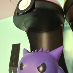 Op de afbeelding: Een paars en zwart verzamelobject in de vorm van een Pok&eacute; Ball met een witte, cirkelvormige knop. Daaronder staat een paarse Gengar-figuur met een brede grijns en puntige oren. De items maken waarschijnlijk deel uit van een grotere collectie.