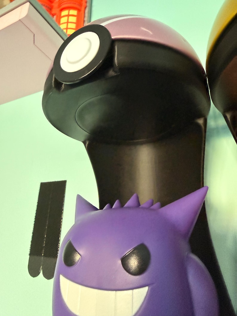 Op de afbeelding: Een paars en zwart verzamelobject in de vorm van een Pok&eacute; Ball met een witte, cirkelvormige knop. Daaronder staat een paarse Gengar-figuur met een brede grijns en puntige oren. De items maken waarschijnlijk deel uit van een grotere collectie.