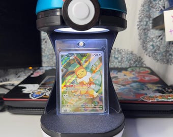May include: A spherical display stand shaped like a red, blue, and white Poke Ball with a black center band. The stand features an integrated LED light that illuminates a vertical trading card encased in a clear protective plastic sleeve.
