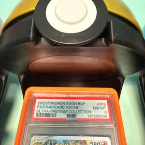 May include: A PSA graded 2022 Pokemon SWSH BSP Charizard VSTAR trading card in a clear plastic slab with an orange border, positioned in front of a gold and black spherical container featuring a central white circular button on its top surface.