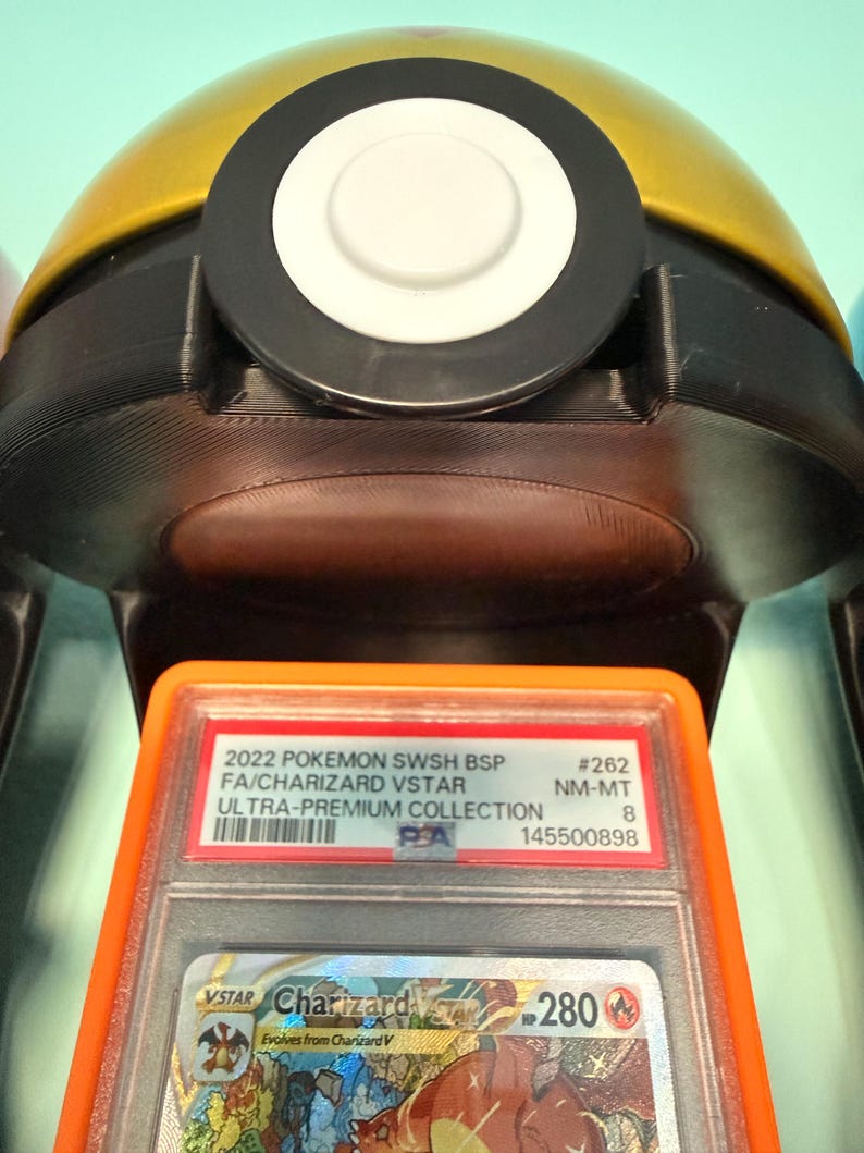 May include: A PSA graded 2022 Pokemon SWSH BSP Charizard VSTAR trading card in a clear plastic slab with an orange border, positioned in front of a gold and black spherical container featuring a central white circular button on its top surface.