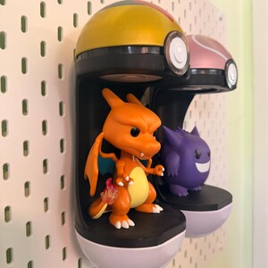 Op de afbeelding: Twee Pok&eacute;mon-thema displayrekken gemonteerd op een wit pegboard. E&eacute;n plank bevat een oranje Charizard-figuur en de andere een paarse Gengar-figuur. De planken zijn zwart met witte bases en de bovenkanten zijn geel en roze.