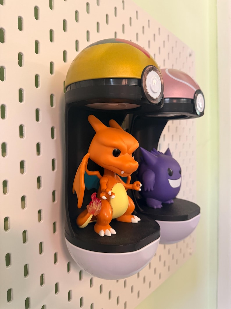 Op de afbeelding: Twee Pok&eacute;mon-thema displayrekken gemonteerd op een wit pegboard. E&eacute;n plank bevat een oranje Charizard-figuur en de andere een paarse Gengar-figuur. De planken zijn zwart met witte bases en de bovenkanten zijn geel en roze.