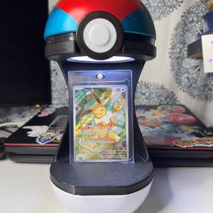 Può includere: Un espositore sferico a forma di Poké Ball rossa, blu e bianca con una fascia centrale nera. Il supporto è dotato di una luce LED integrata che illumina una carta collezionabile verticale inserita in una custodia protettiva trasparente.