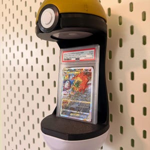 May include: A yellow and white spherical display stand shaped like a Poke Ball, featuring a black center section that holds a graded Charizard VSTAR trading card in a clear plastic PSA slab. The card displays colorful artwork and text.