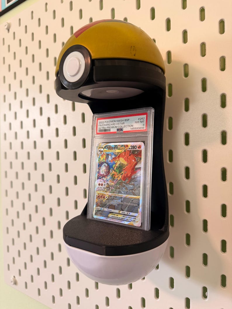 May include: A yellow and white spherical display stand shaped like a Poke Ball, featuring a black center section that holds a graded Charizard VSTAR trading card in a clear plastic PSA slab. The card displays colorful artwork and text.