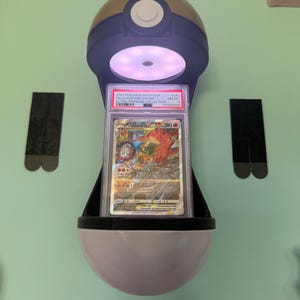 May include: A Pokémon card in a protective case, displayed in a Poké Ball-shaped holder. The card features a Charizard VSTAR illustration. The holder is black, white, and gold, with a built-in light. The card is graded and labeled with details.