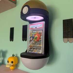 May include: A wall-mounted display stand shaped like a gold and black Poke Ball with an integrated light illuminating a graded Charizard VSTAR trading card in a clear plastic slab. The card features a red dragon illustration and text.