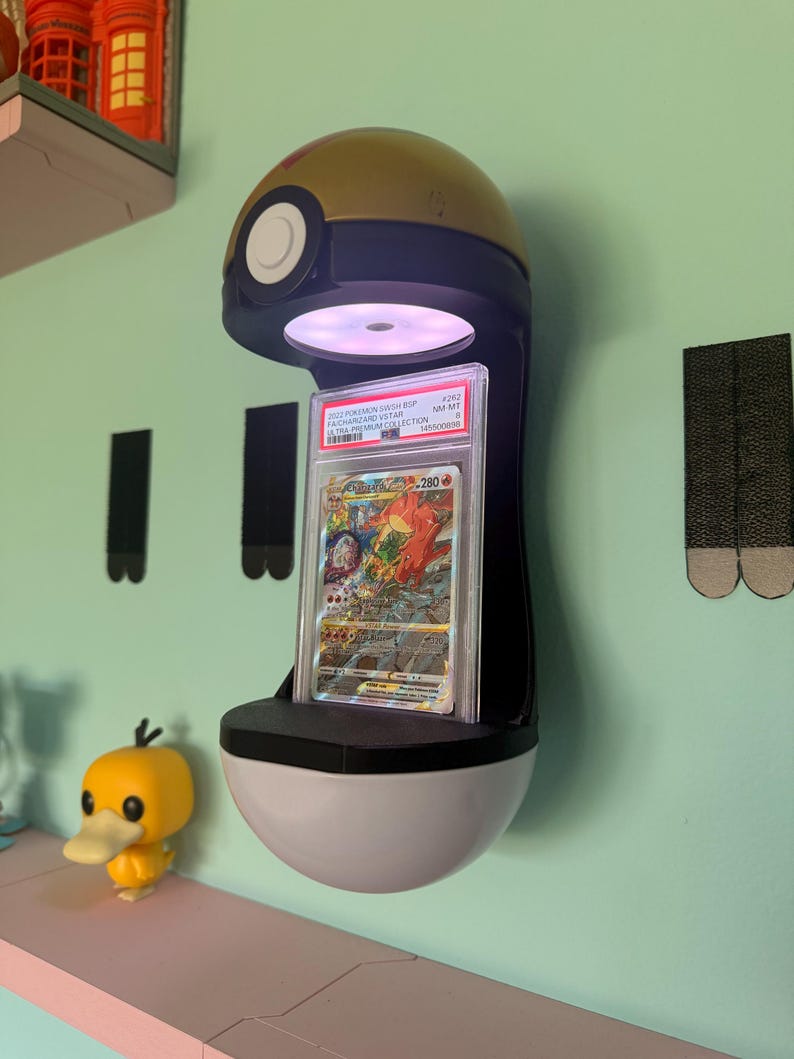May include: A wall-mounted display stand shaped like a gold and black Poke Ball with an integrated light illuminating a graded Charizard VSTAR trading card in a clear plastic slab. The card features a red dragon illustration and text.