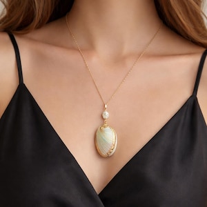 Puede incluir: Collar dorado con un colgante en forma de concha. El colgante ovalado presenta un patrón verde claro y blanco con detalles dorados. Una pequeña perla redonda se encuentra encima del colgante, sobre una blusa negra.