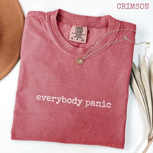 Könnte beinhalten: Karmesinrotes T-Shirt mit dem weißen Schriftzug „everybody panic“. Das Shirt hat einen Rundhalsausschnitt und ein kleines Etikett am Ausschnitt. Eine silberne Halskette mit Anhänger liegt über dem Shirt.