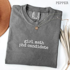 May include: A gray Comfort Colors t-shirt with the text "girl math phd candidate" in white. A gold-colored necklace with a pendant is draped across the shirt. The shirt is folded and displayed on a white surface.