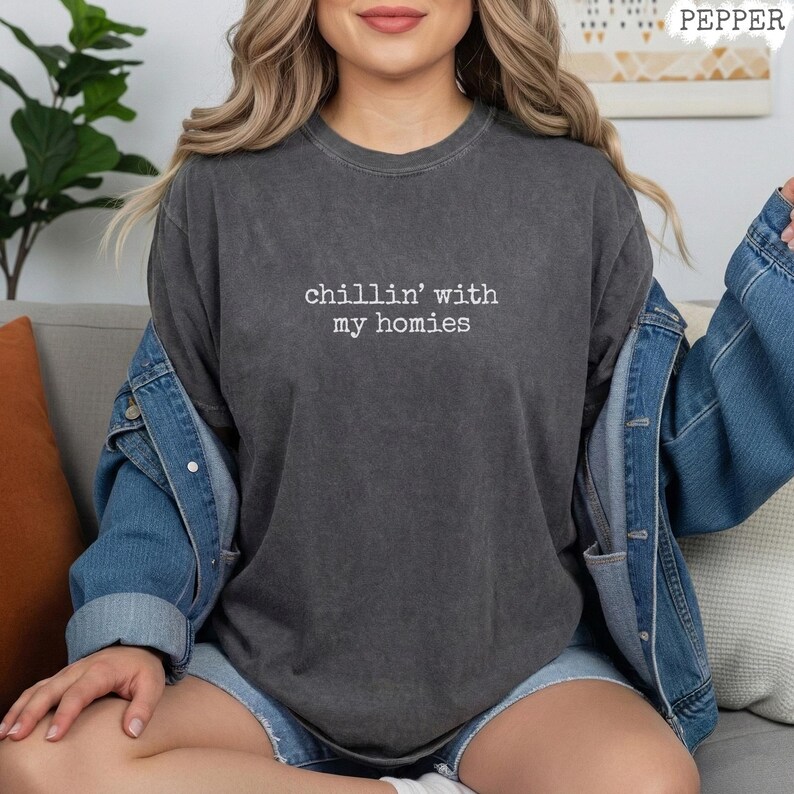 以下が含まれることがあります： 「chillin' with my homies」の白い文字が入った濃いグレーのTシャツ。デニムジャケットとショートパンツと合わせています。全体的にカジュアルでリラックスしたスタイルです。