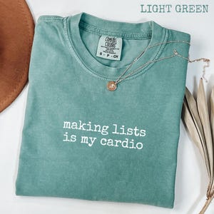 May include: A light green short-sleeve t-shirt with a crew neckline and the white text "making lists is my cardio" printed on the front. The shirt features a Comfort Colors brand label at the inner neck and a thin gold-tone chain necklace.