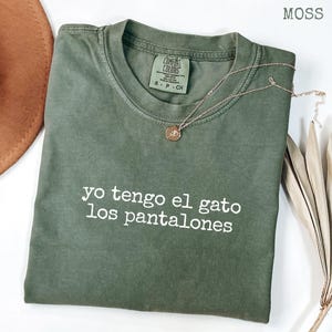 May include: A moss green t-shirt with the phrase "yo tengo el gato los pantalones" in white lettering. A gold-colored necklace with a pendant is draped across the shirt. The shirt is folded neatly on a white surface.