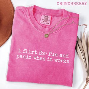 Może przedstawiać: Różowa koszulka Comfort Colors z białym napisem "i flirt for fun and panic when it works". Złoty naszyjnik z wisiorkiem jest zarzucony na koszulkę. Słowo "CRUNCHBERRY" znajduje się na górze.