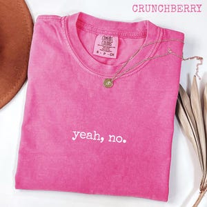 Könnte beinhalten: Ein leuchtend pinkfarbenes T-Shirt mit dem weißen Schriftzug "yeah, no." Das Shirt hat einen Rundhalsausschnitt und ein kleines Etikett. Eine goldene Halskette mit Anhänger liegt auf dem Shirt. Das Wort "CRUNCHBERRY" steht oben.