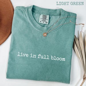 May include: A light green t-shirt with the words "live in full bloom" printed in white. A silver necklace with a round pendant is on the shirt. The shirt is folded and has a Comfort Colors tag.