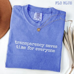 Puede incluir: Una camiseta de manga corta en color azul Flo con cuello redondo y etiqueta Comfort Colors en el cuello interior. La prenda presenta el texto blanco "transparency saves time for everyone" impreso en el pecho y un collar con colgante dorado.