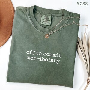 Könnte beinhalten: Ein moosgrünes T-Shirt mit dem weißen Schriftzug "off to commit mom-foolery". Eine goldfarbene Halskette mit Anhänger liegt auf dem Shirt. Das Shirt ist auf einer weißen Oberfläche gefaltet.