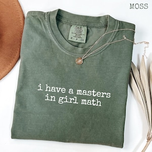 Può includere: Una maglietta verde muschio con la scritta bianca "i have a masters in girl math". Una collana d'oro con un ciondolo è drappeggiata sulla maglietta. La maglietta è piegata su una superficie bianca, con un cappello marrone e una pianta secca sullo sfondo.