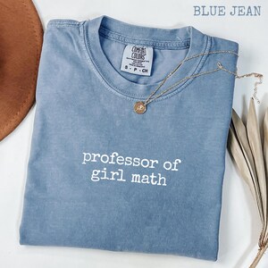 Può includere: Una maglietta blu jeans con la scritta bianca "professor of girl math". La maglietta è piegata e ha una collana d'oro con un ciondolo. L'etichetta del marchio "Comfort Colors" è visibile. Un cappello marrone e steli di piante secche sono sullo sfondo.