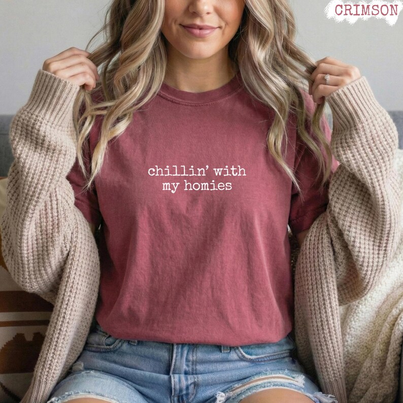 以下が含まれることがあります： 「chillin' with my homies」の白い文字が入ったマルーン色のTシャツ。デニムショーツとベージュのカーディガンを合わせています。右上には「CRIMSON」の文字があります。