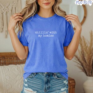 以下が含まれることがあります： 白い文字で「chillin' with my homies」と書かれた青いTシャツ。Tシャツは無地で半袖です。モデルは青いデニムショーツを着用しています。