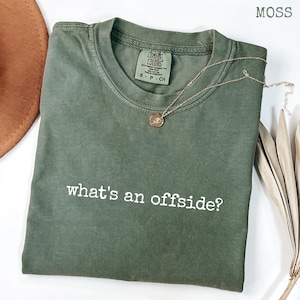 Puede incluir: Una camiseta verde musgo con el texto "what's an offside?" en blanco. La camiseta tiene cuello redondo y mangas cortas. Un collar dorado con un colgante está drapeado sobre la camiseta.