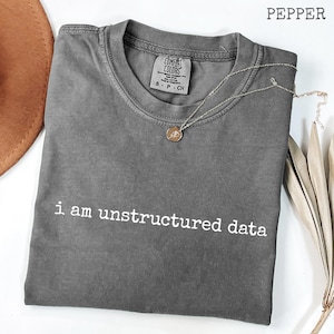 May include: A gray, short-sleeved t-shirt with the text "i am unstructured data" in white. A gold-colored necklace with a pendant is draped across the shirt. The shirt is folded on a white surface.