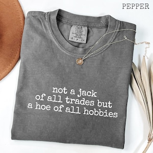 Peut inclure: Un t-shirt à manches courtes de couleur poivre avec une encolure ronde. Le t-shirt présente un texte blanc qui dit : not a jack of all trades but a hoe of all hobbies. Un collier doré avec un petit pendentif circulaire repose sur le col.