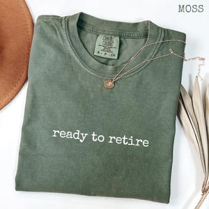 May include: A moss green crew neck t-shirt with the text ready to retire printed in white across the chest. The shirt features a Comfort Colors brand label at the inner neckline and is paired with a thin gold chain necklace with a small round pendant.