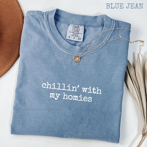 以下が含まれることがあります： 「chillin' with my homies」の白い文字が入った、ブルーデニムのComfort Colors Tシャツ。ペンダント付きのゴールドネックレスがシャツの上にあります。茶色の帽子と乾燥した植物が背景にあります。