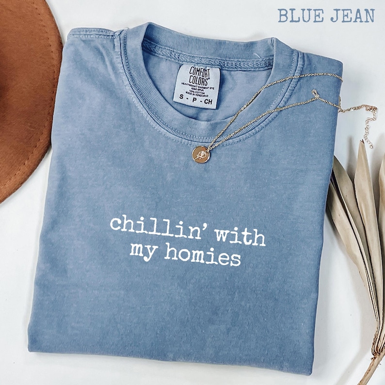 以下が含まれることがあります： 「chillin' with my homies」の白い文字が入った、ブルーデニムのComfort Colors Tシャツ。ペンダント付きのゴールドネックレスがシャツの上にあります。茶色の帽子と乾燥した植物が背景にあります。
