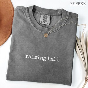Könnte beinhalten: Ein graues T-Shirt mit kurzen Ärmeln und dem weißen Aufdruck "raising hell". Eine goldene Halskette mit Anhänger liegt auf dem Shirt. Das Shirt ist auf einer weißen Oberfläche gefaltet.