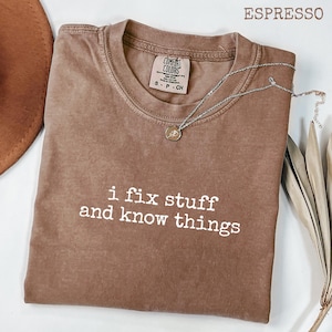 May include: Espresso-colored t-shirt with the phrase "i fix stuff and know things" in white lettering. The shirt has a crew neck and a Comfort Colors tag. A silver necklace with a pendant is draped across the shirt.