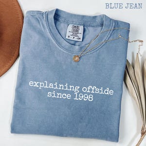 Puede incluir: Una camiseta Comfort Colors azul vaquero con el texto "explaining offside since 1998" en blanco. Un collar dorado con un colgante descansa sobre la camiseta. La camiseta está doblada y colocada sobre una superficie blanca.
